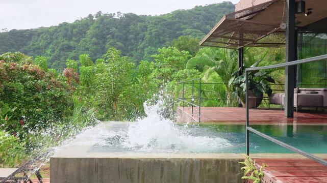 Young boy jumps into a concrete infinity pool at a luxurious modern tropical villa surrounded by lush green rainforest mountains, wooden deck and contemporary architecture visible on overcast day.
