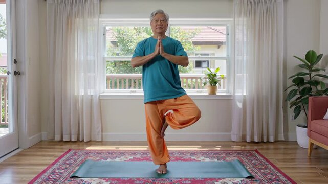 Senior man practicing yoga tree pose at home. Asian man balancing on one leg in living room. Healthy lifestyle for elderly, wellness and physical fitness exercise for mental relaxation and body