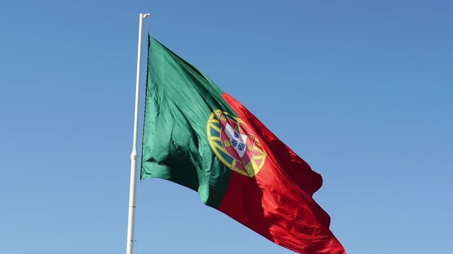 Big flag of Portugal fluttering in the wind on a white flagpole. Captured during a bright sunny day near Edward VII Park in Lisbon. Medium low angle shot 