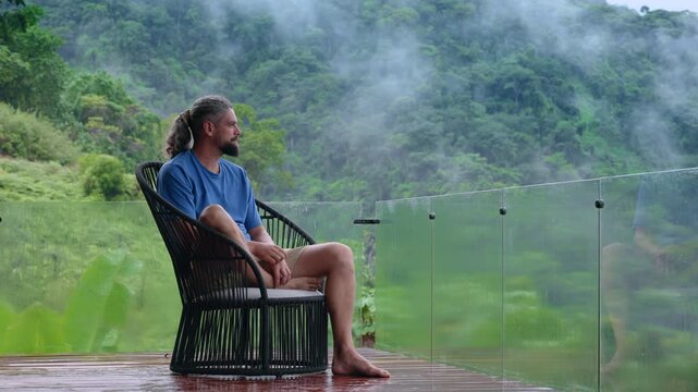 Relaxed bearded man with long hair tied back sits barefoot in modern black wicker chair on rain-soaked wooden deck with glass balustrade, gazing at misty green jungle-covered mountains and fog.