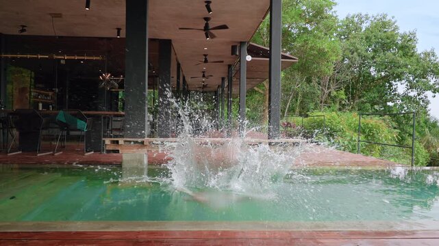 Young boy leaps into turquoise infinity pool at modern tropical villa with wooden deck, ceiling fans overhead, lush green jungle surroundings, and contemporary open-air architecture on cloudy day.
