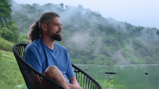 Bearded man with long hair tied in ponytail relaxes in modern wicker chair on balcony overlooking misty tropical rainforest mountains.