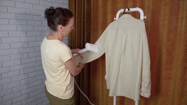Woman steaming clothes with handheld garment steamer. Hardworking housewife using a portable vertical garment steamer to iron a wrinkled shirt hanging on a rack