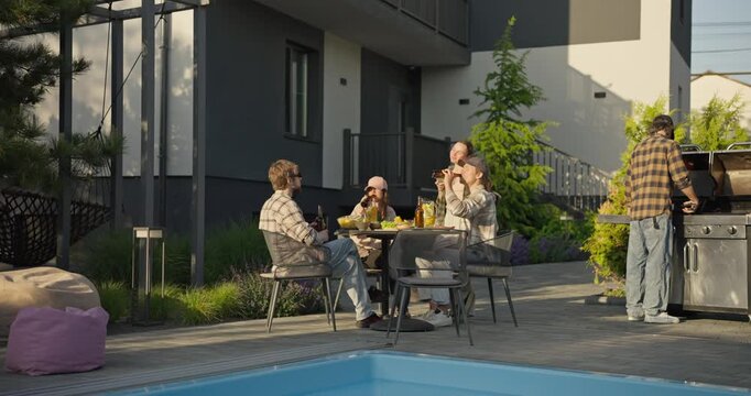 Friends gathering in a backyard by the pool with barbecue grilling in warm daylight