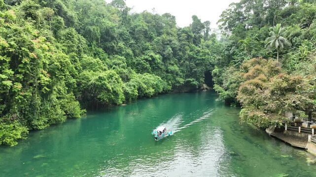 Aerial view of a small boat navigating the emerald green waters of the Green Canyon river surrounded by dense tropical rainforest in Pangandaran Regency, West Java, Indonesia.