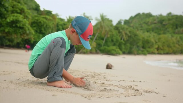 Young boy wearing green shirt and colorful baseball cap crouches barefoot on sandy tropical beach building with sand. Palm trees and lush jungle vegetation line the shore in the background.
