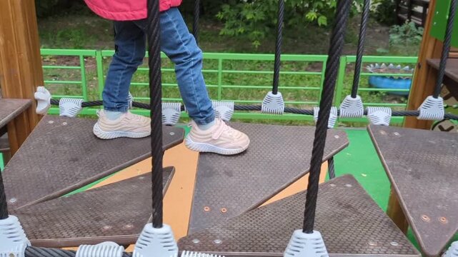 child wearing blue jeans and beige sneakers as they step across textured, triangular platform steps on a modern playground bridge structure supported by thick black vertical ropes