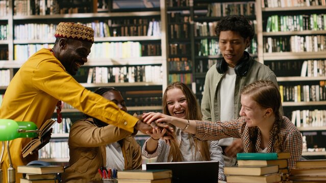 Smiling diverse students stacking hands together in unity at library table, celebrating teamwork and successful group study session, classmates united in academic work for office teamwork themes.
