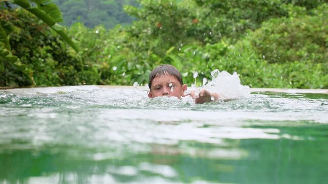 Slow motion footage of a young boy swimming in a tropical infinity pool surrounded by lush green jungle foliage. Water splashes dramatically as child moves through the turquoise water on a cloudy day.