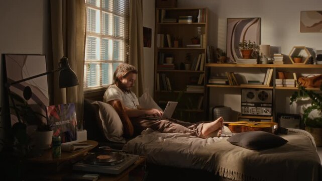 Caucasian young adult man sitting comfortably on bed at home and typing on laptop while writing computer code. For promoting remote work, freelance lifestyle, and flexible hybrid career options