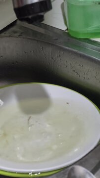 Close up of a little child in pineapple pajamas washing green plates in the kitchen sink diligently.