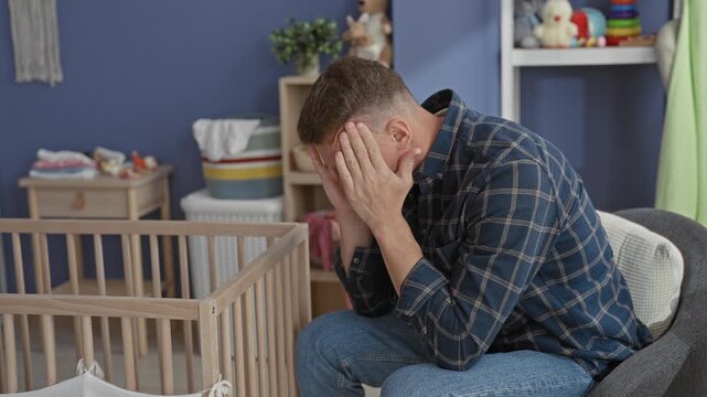 Man seated by crib in nursery with hands covering face and head bowed, toys on shelf and empty cradle visible in building; parenting stress despair.