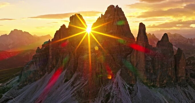Aerial drone view of Tre Cime di Lavaredo at sunrise with dramatic sunstar between the iconic peaks. Golden morning light illuminates the rugged Dolomite landscape in northern Italy.