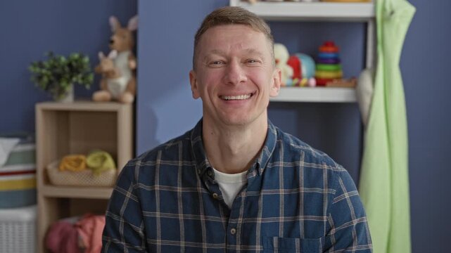 Man smiling with visible teeth in a nursery set inside a studio, wearing a blue flannel shirt and making eye contact; parenthood warmth joy.