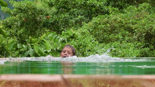 Child splashing and swimming in an infinity pool surrounded by dense tropical foliage. Water droplets spray as the boy moves through the clear green water with jungle trees behind him.