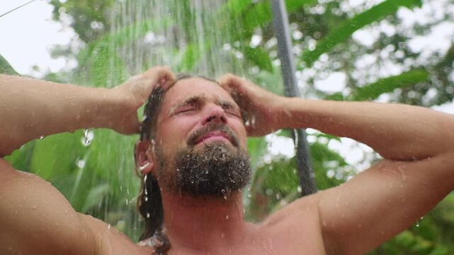 Slow motion close-up of bearded man washing his long hair under outdoor shower surrounded by lush tropical greenery. Water droplets cascade down as he runs hands through wet hair with eyes closed.