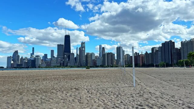 A wide view shows a beach in Chicago with volleyball nets set up in the sand. The skyline of tall buildings rises in the background under a bright sky.