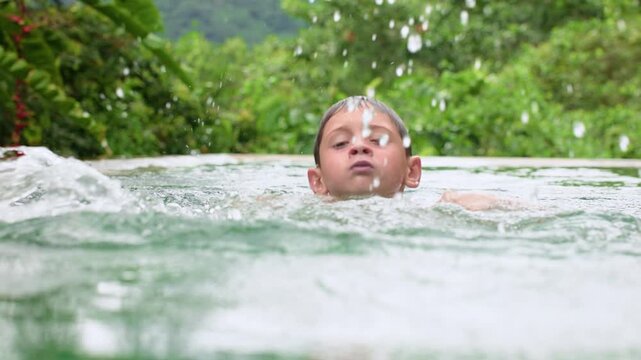 Slow motion footage of young boy swimming in a tropical outdoor pool surrounded by lush green foliage. Water droplets splash dramatically as child moves through turquoise water during summer vacation.