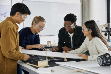 Fototapeta premium Group of young people wearing safety glasses working together on an aircraft model in a bright engineering classroom or lab.