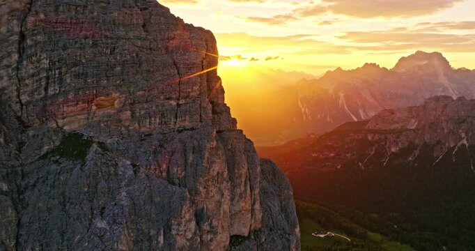 Smooth aerial flight along towering Dolomite cliffs during sunset. Golden sunlight creates a dramatic sunstar effect, illuminating rugged rock formations and alpine landscapes in Italy.