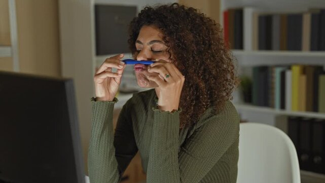 Young hispanic woman balancing a blue pen on her upper lip with hand raised near a computer monitor in an office building; playful distraction.