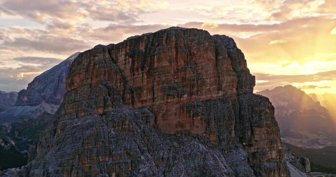 Smooth aerial flight along towering Dolomite cliffs during sunset. Golden sunlight creates a dramatic sunstar effect, illuminating rugged rock formations and alpine landscapes in Italy.