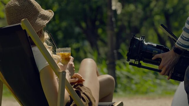 Woman sitting on a lounge chair in an outdoor cafe. Holding an iced coffee with a straw. Man with a film camera shooting a film