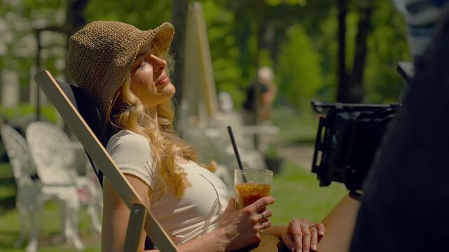 Woman sitting on a lounge chair in an outdoor cafe. Holding an iced coffee or tea with a straw. Man with a film camera shooting a film