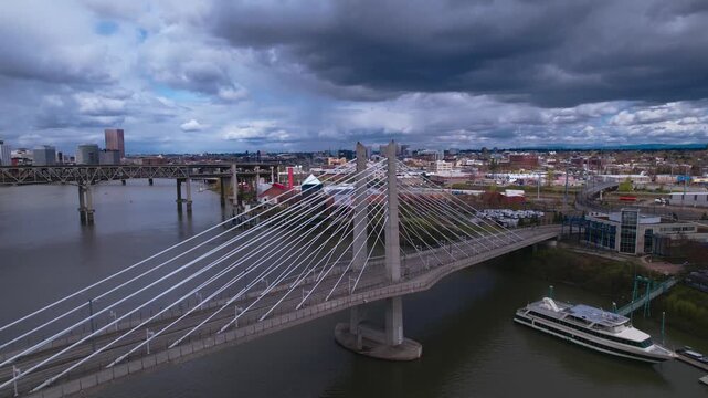 Aerial panoramic view of the Tilikum Crossing bridge spanning the Willamette River in Portland, Oregon, featuring the urban cityscape under a dramatic cloudy sky with river boats docked