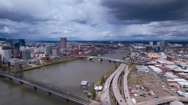 Wide aerial panorama of the Portland city skyline and Willamette River under a dramatic cloudy sky, featuring major bridges and highway traffic flowing through the urban landscape in Oregon, USA
