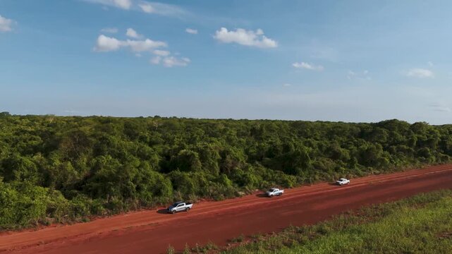 Aerial view of a convoy of white pickup trucks traveling along a winding red dirt road through a dense green tropical forest under a clear blue sky with scattered clouds in a rural landscape