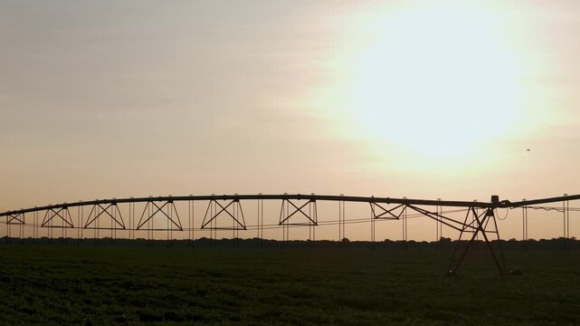 A large center pivot irrigation system stands in a lush green field under a bright setting sun, showcasing modern agricultural technology and water management in a rural landscape at dusk