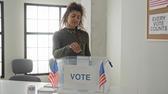 Woman drops ballot into transparent box at voting building with small american flags on desk and voter sticker roll visible; civic duty pride.