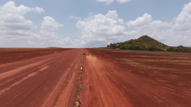 A wide aerial view of a vehicle driving along a long red dirt road in a remote arid landscape with a small green hill and a blue cloudy sky in the background, kicking up a trail of dust