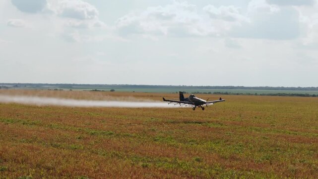 Small agricultural airplane flying at low altitude over a vast rural field while spraying pesticides or fertilizers, creating a visible white trail against a cloudy sky background