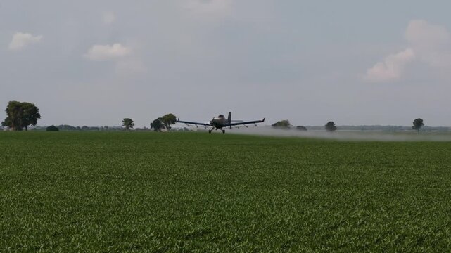 A specialized crop duster airplane flies at a very low altitude over a lush green agricultural field, releasing a fine mist of liquid fertilizer or pesticide under a vast, overcast sky