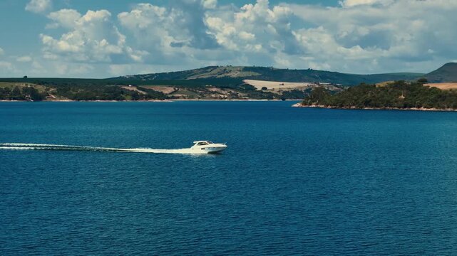 A sleek white motorboat speeds across the deep blue waters of a scenic lake, leaving a white wake behind it under a bright cloudy sky with rolling green hills in the distant background