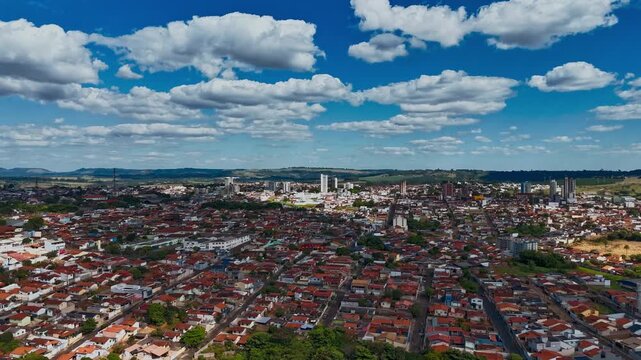 Wide aerial panoramic view of a sprawling urban city with red tiled roofs under a bright blue sky filled with white fluffy clouds, showcasing the dense residential architecture and green spaces