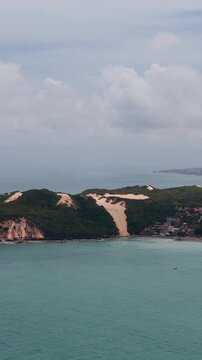 Aerial panoramic view of the iconic Morro do Careca sand dune at Ponta Negra beach in Natal, Brazil, featuring lush green vegetation, turquoise ocean waters, and a coastal village under a cloudy sky