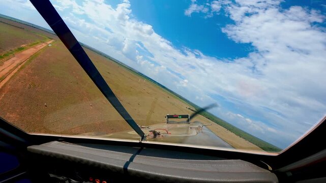 Wide-angle view from a helicopter cockpit flying over rural agricultural fields under a bright blue sky with white clouds, showing the dashboard and windshield during a low-altitude flight