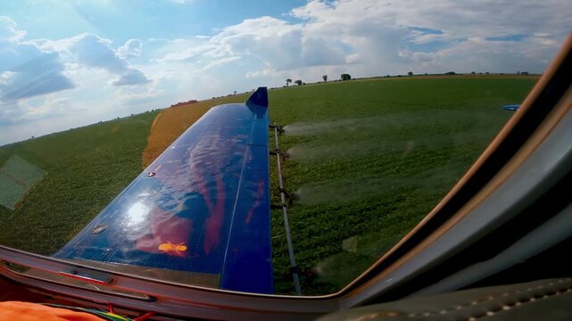 View from the cockpit of a crop duster aircraft flying low over vast green agricultural fields, spraying liquid fertilizers or pesticides from its wings under a bright summer sky with clouds