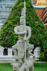 A stone guardian statue stands guard in front of a lush green tree at The Grand Palace, Bangkok, Thailand. The intricate details of the statue depict a mythical warrior. © Zenstratus