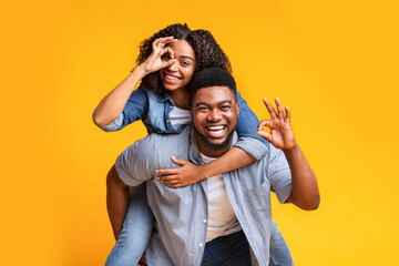 Fototapeta premium Smiling Black Young Couple Enjoying Playful Moment. Man Giving Woman Piggyback Ride While Showing Hand Gestures, Having Fun Together In Studio At Modern Yellow Background. Concept Of Happiness.