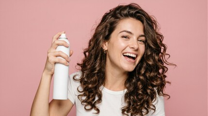 Fototapeta premium Happy woman with curly hair holding a hairspray bottle, isolated on a pink background