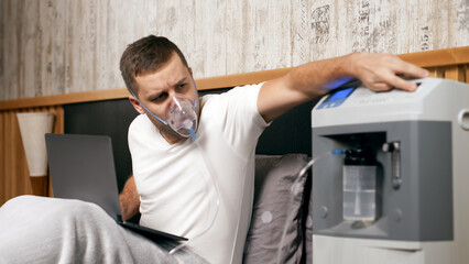 Fototapeta premium Modern oxygen concentrator used by a man wearing a nasal cannula while sitting on a bed with a laptop, in a cozy indoor setting with wooden accents