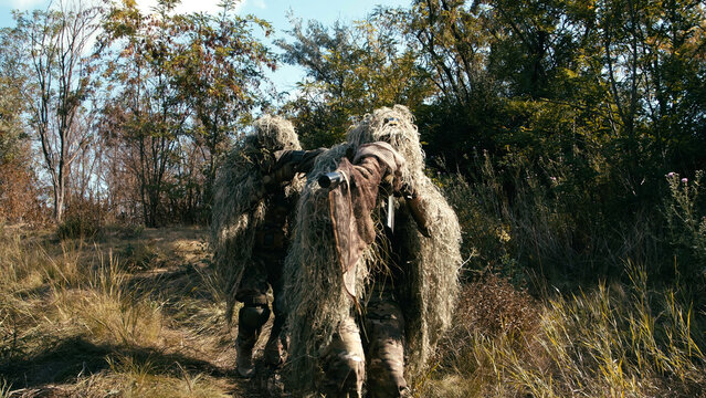 Sniper team in camouflage gear carrying tactical equipment through dense foliage in a forested area during a covert mission in a natural environment
