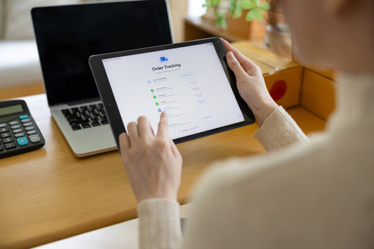 Person tracking an online order on a tablet at a home workspace, with laptop, calculator, and shipping boxes on the desk