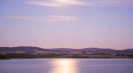 Fototapeta premium A tranquil lake reflects the soft hues of a twilight sky, with rolling hills and distant lights creating a peaceful landscape.