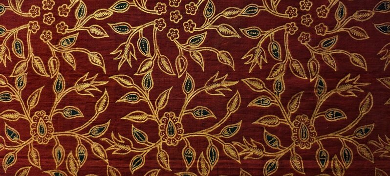 Richly patterned batik fabric featuring intricate golden floral embroidery on a deep red background. The design showcases repeating motifs of stylized flowers, leaves, and vines, with dark blue detail