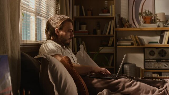 Caucasian young male programmer or software developer sitting in bed, typing on laptop computer at home. Video represents remote work, freelance lifestyle, and flexible working hours
